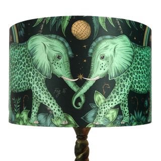 The Large Teal Zambezi Silk Lampshade features two enchanting Zambezi elephants on the front intertwining trunks, with vibrant greens and golds on a deep Navy background